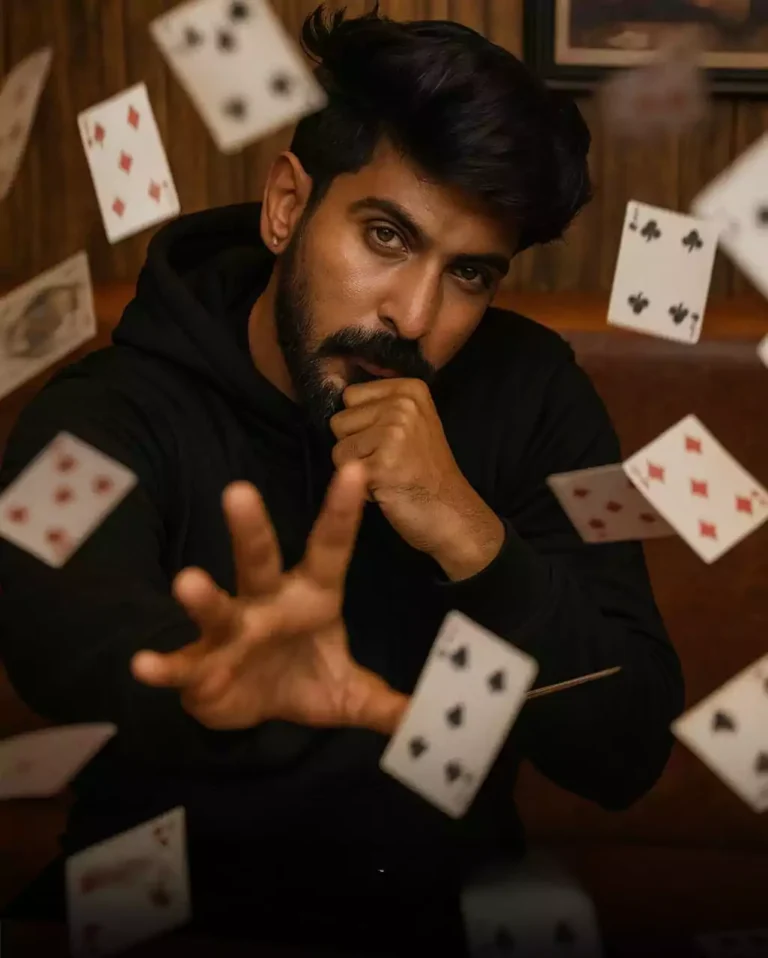 A young man with dark hair(maintain exact face and skin tone with sharp features) wearing a black hoodie sits in a rustic wooden booth, staring intensely at the camera with one hand supporting his chin and the other reaching forward as if performing telekinesis. Dozens of playing cards fly around him in mid-air, captured n dynamic slow-motion with motion blur. The background shows vertical wooden panels and a framed photo of a man playing cards.

