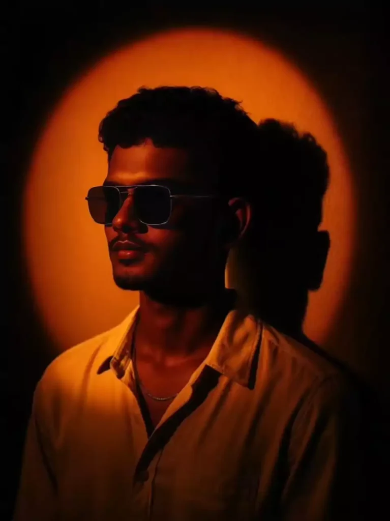 Create a cinematic studio portrait of the uploaded person, with a dramatic orange-yellow spotlight forming a glowing circular halo on the wall behind them. The lighting should cast deep shadows on the wall, especially the person's head shadow to the right side. The person should have short curly or textured hair, wearing a cream or off-white open-collar shirt and a thin chain necklace. The subject is facing slightly left with a soft, introspective expression. The background is minimalist and dark, allowing the warm lighting to dominate the scene. Capture a soft, moody, golden glow across the face and upper chest, blending realism with an artistic sunset tone."

